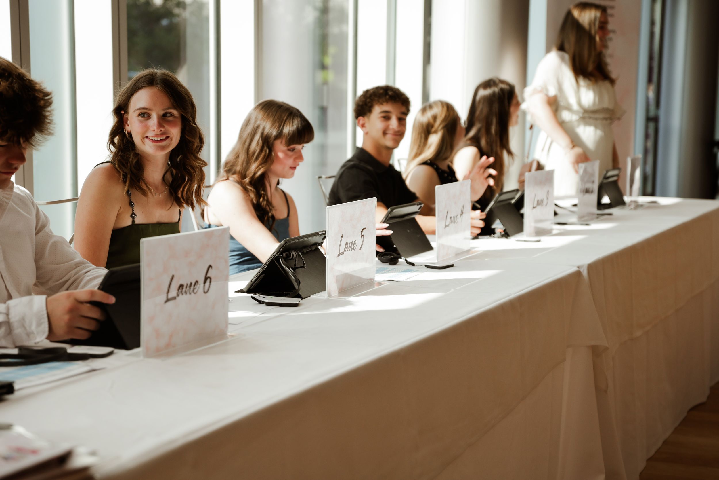 Young guests smile while seated at a long registration table during a bright and modern event venue gathering.