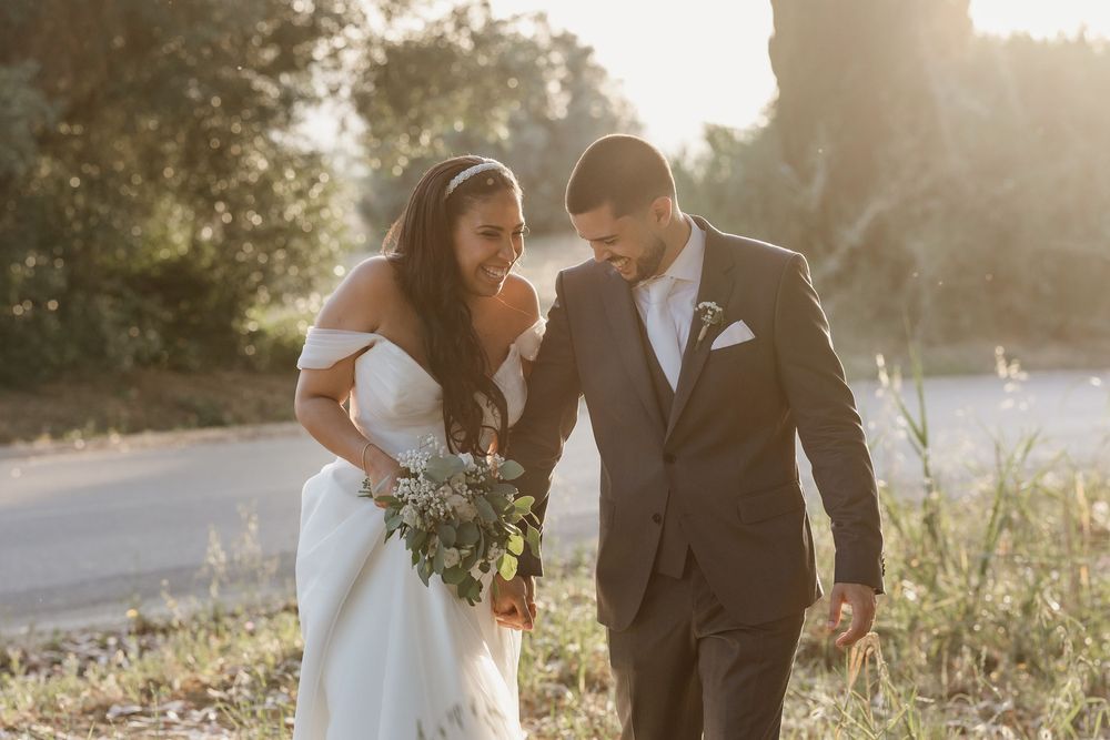 A couple in wedding attire walks together through a sunlit meadow during golden hour.