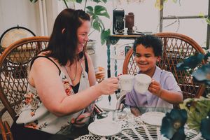 Woman and child clinking mugs together at a table, smiling and enjoying a tea party indoors.
