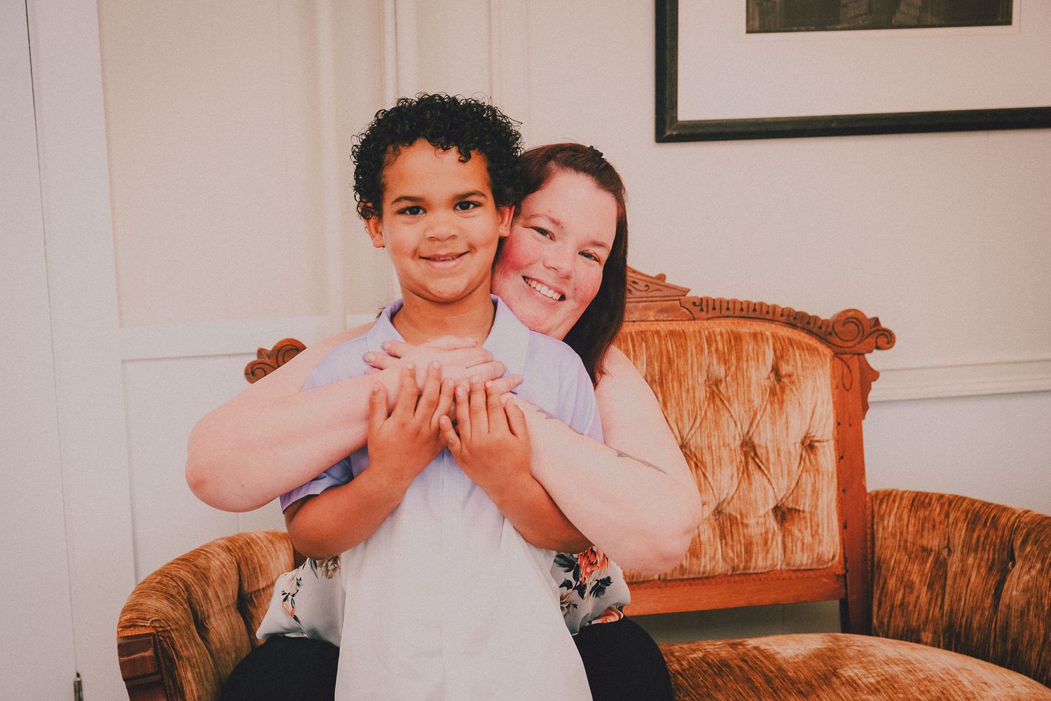 A woman hugs a young boy from behind while sitting on an antique sofa, both smiling warmly indoors.