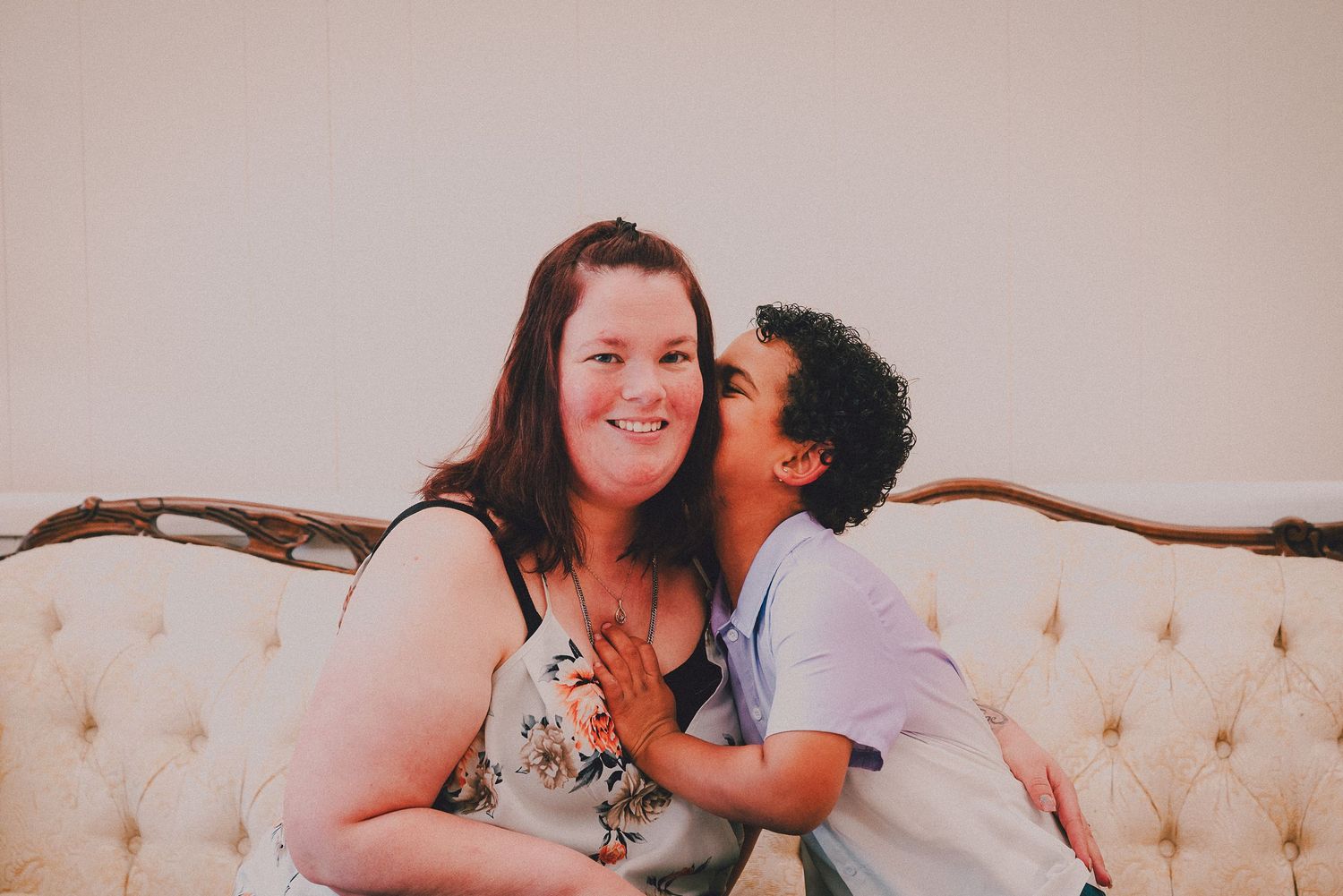 A woman smiles as a child kisses her cheek while sitting on a tufted white vintage sofa.