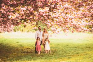 A family stands beneath blooming pink cherry blossoms in a sunlit spring meadow.