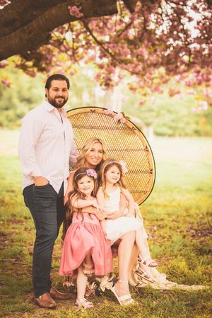 A family sits together on a peacock chair under blooming cherry blossoms in a sunlit spring garden setting.