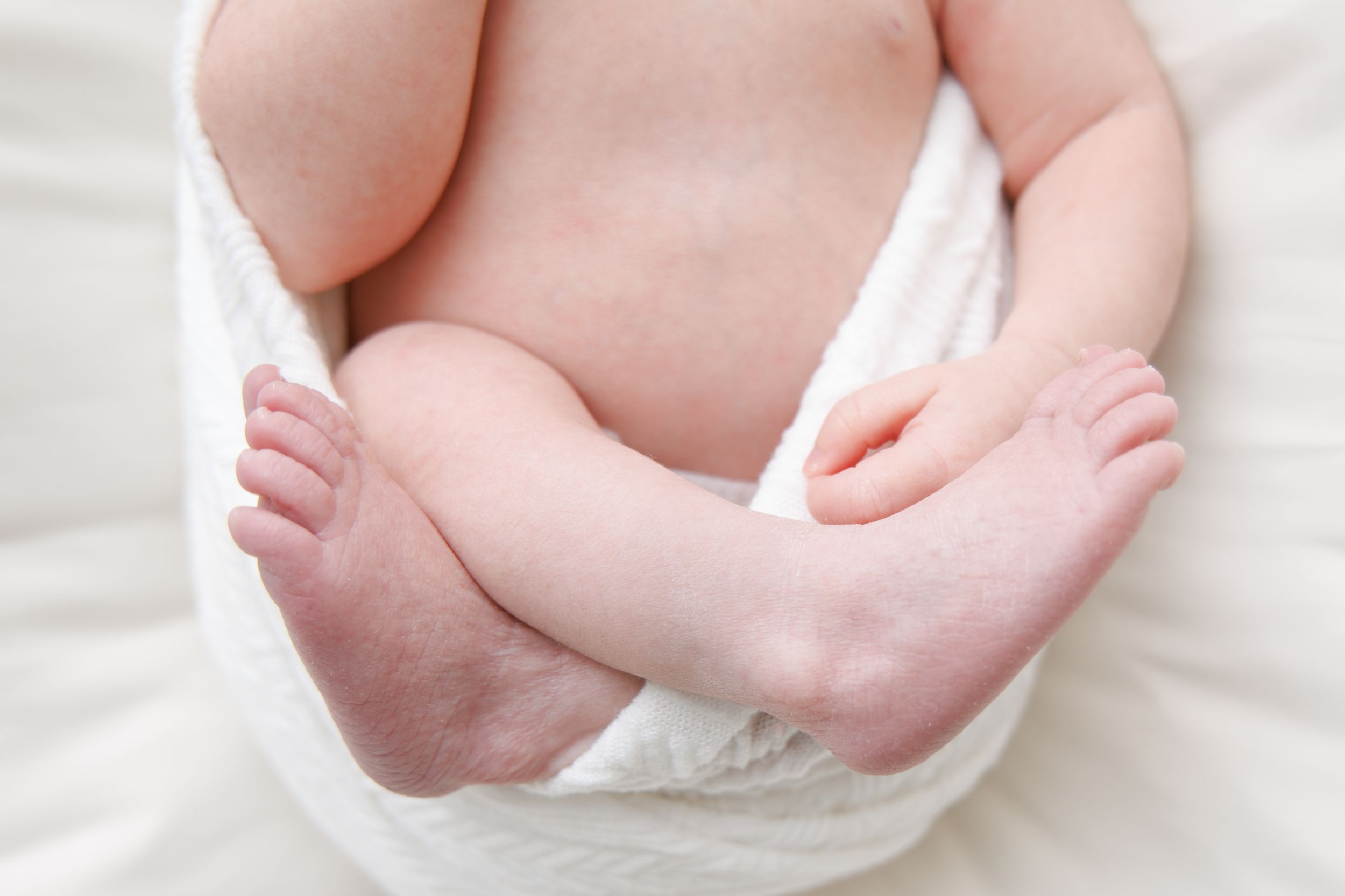 Close up of tiny baby feet and chubby legs wearing a white diaper against white bedding.