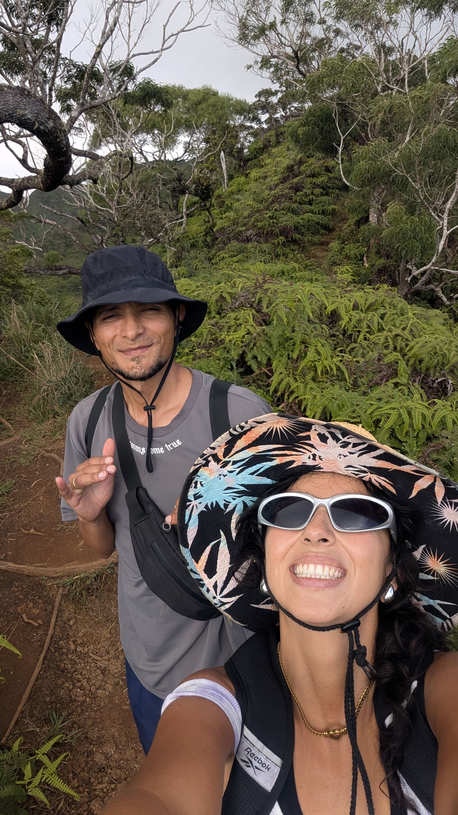 Two hikers wearing bucket hats smile for a selfie on a lush green tropical hiking trail.