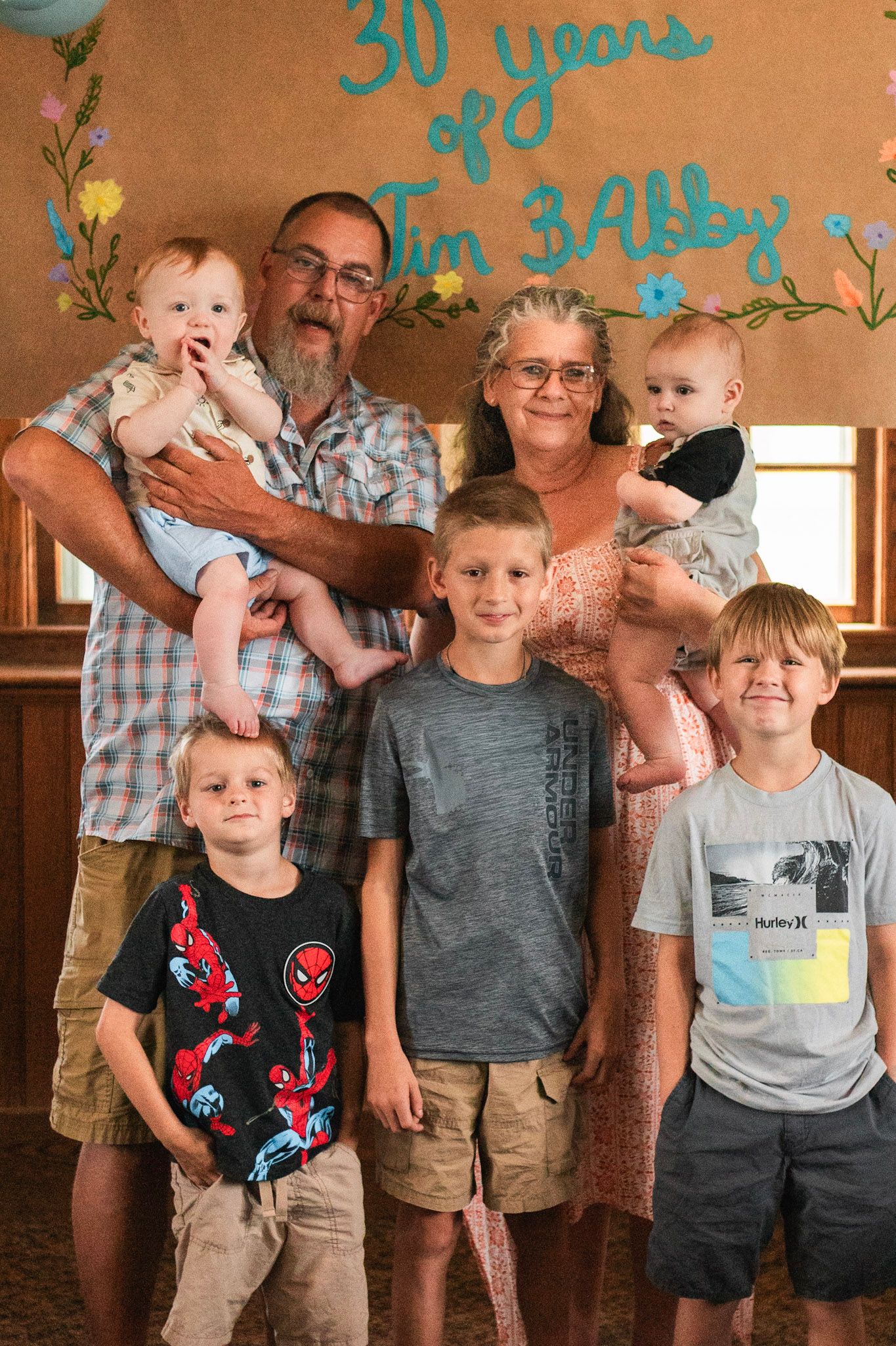 An older couple celebrates 30 years together surrounded by four young boys and two babies indoors.