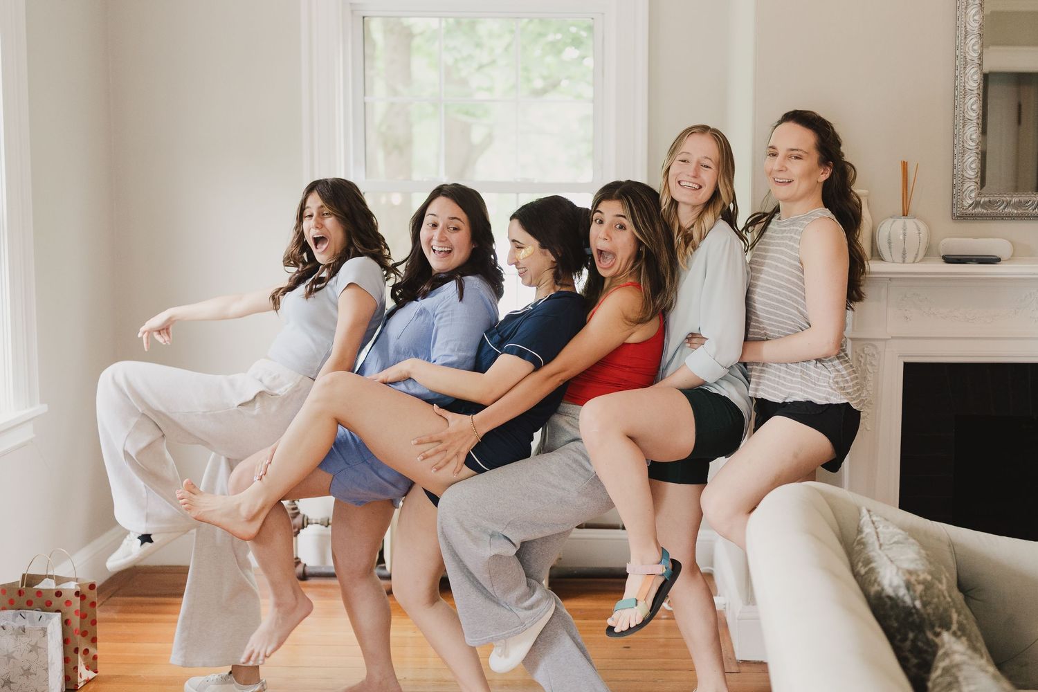 Six women sitting together laughing and posing playfully in a bright living room during a fun group photo session.