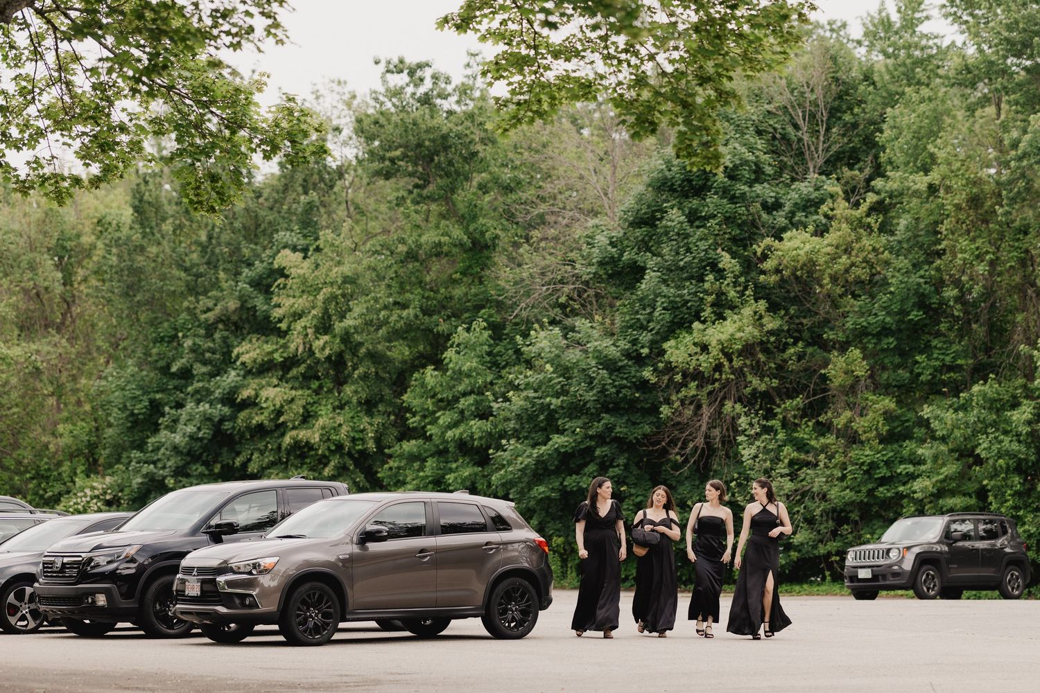 Four bridesmaids in black gowns pose in a parking lot surrounded by cars and lush green trees.