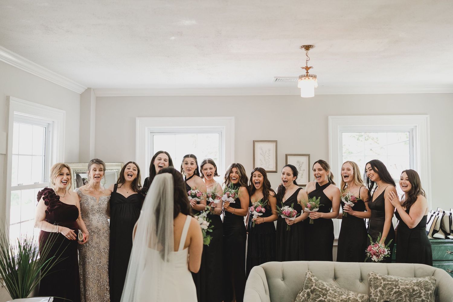 Bridesmaids in black dresses react with joy as bride in white veil reveals her wedding look indoors.