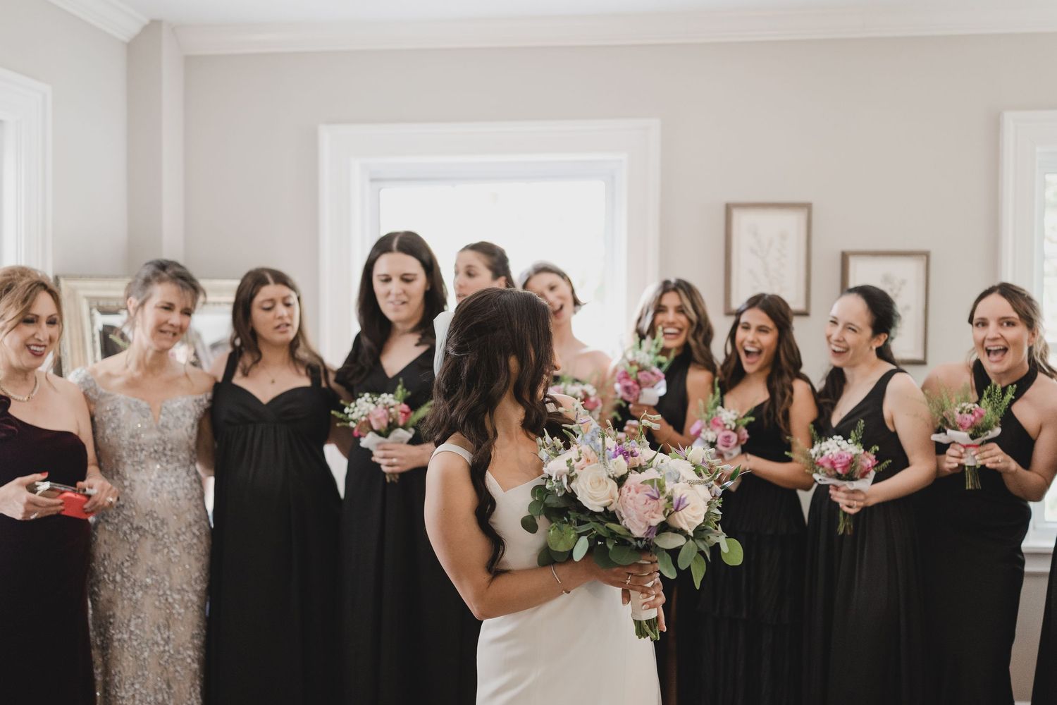 Bride reveals wedding dress to bridesmaids in black gowns holding bouquets in elegant white room.