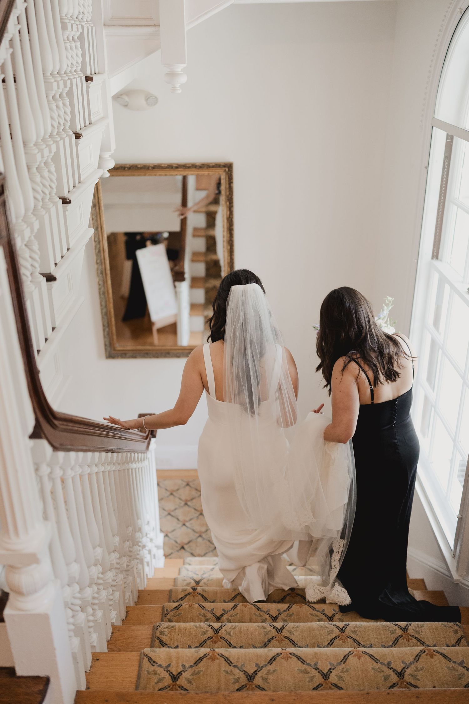 Bride in white gown and veil walks up elegant staircase assisted by bridesmaid in black dress.