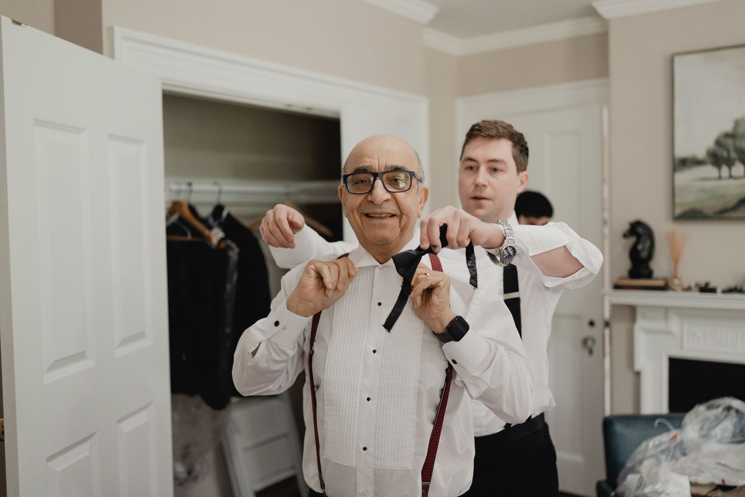 Groom helps older man adjust suspenders while getting ready in bright dressing room before wedding.