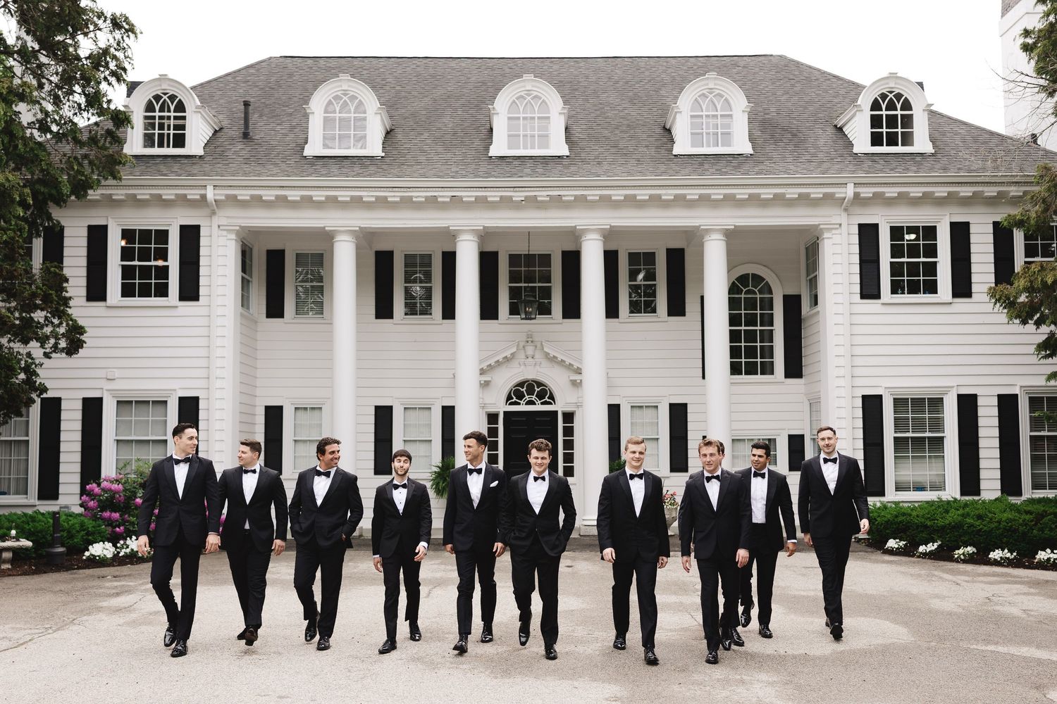 Groomsmen in black tuxedos walking in front of a grand white colonial mansion with black shutters.