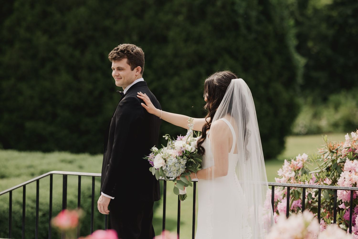 Bride in white gown and veil taps groom's shoulder during first look moment, surrounded by lush greenery and pink flowers.