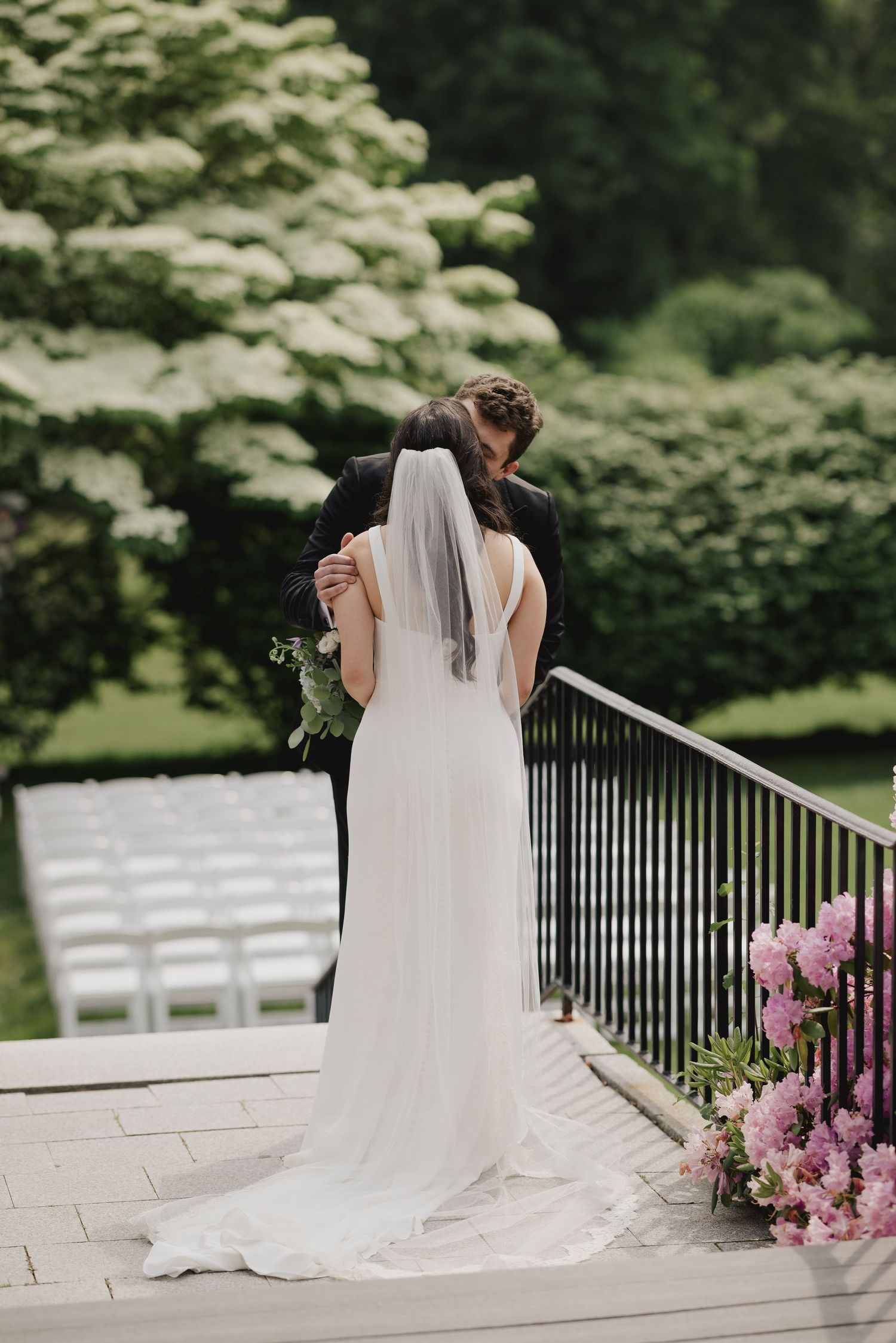 Bride and groom share intimate moment on garden terrace with lush greenery and white ceremony chairs in background.