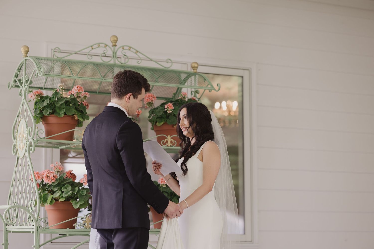 Bride and groom exchange vows outdoors near a vintage green iron plant stand with terracotta pots of pink flowers.