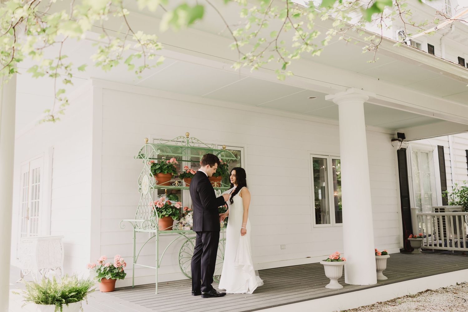 Bride and groom share a romantic moment on a white columned porch adorned with potted plants at an elegant wedding venue.
