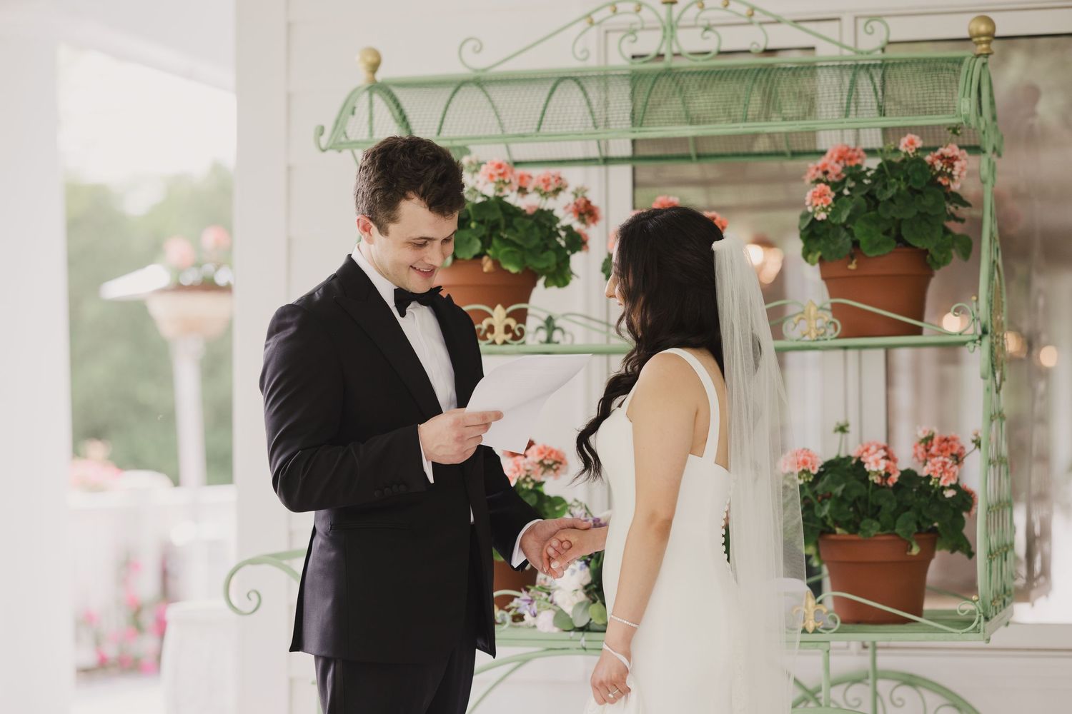 Groom reads vows to bride on decorated porch with vintage green plant stand and pink flowers.