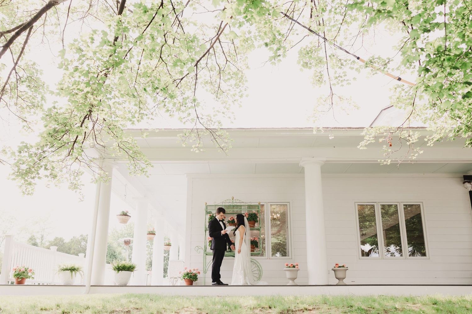 Bride and groom share romantic moment outside classic white columned venue framed by green trees.