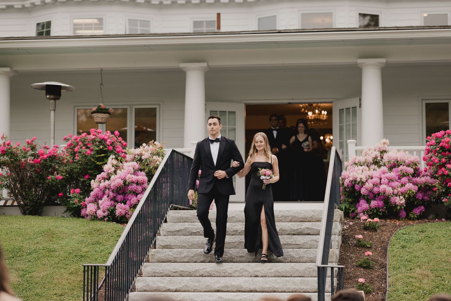 Elegant couple descends grand white mansion steps surrounded by vibrant pink rhododendrons at formal wedding event.