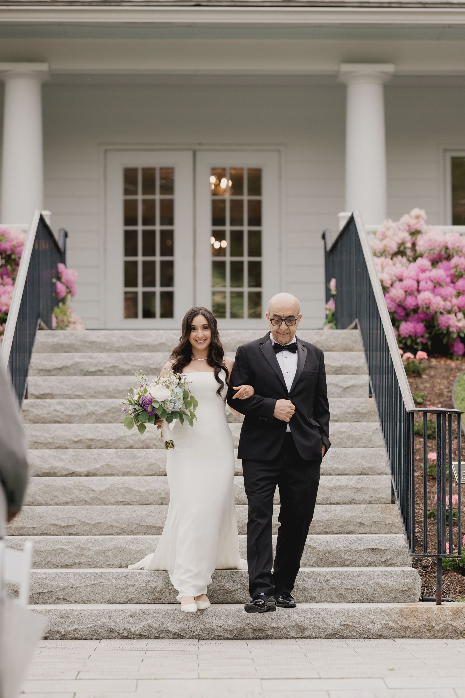 Bride walks down stone steps with escort beside elegant white venue surrounded by blooming pink flowers.