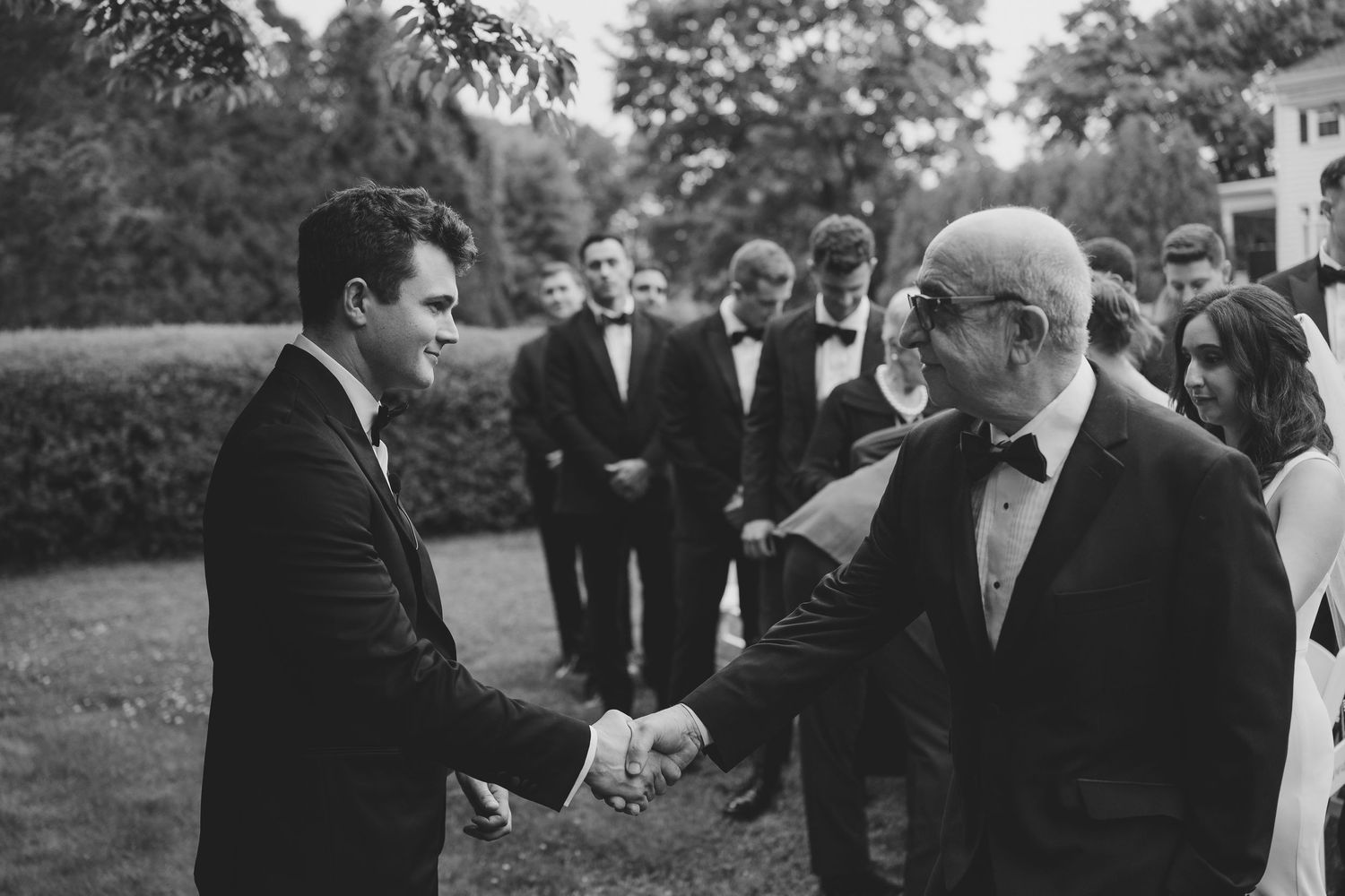 Groom in tuxedo shakes hands with older gentleman at outdoor wedding ceremony in black and white photo.