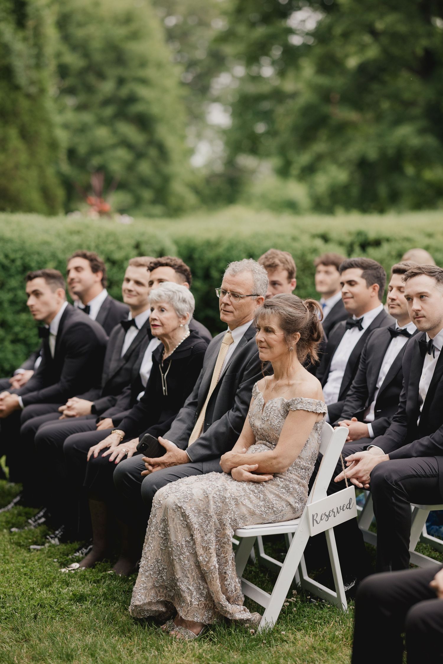 Wedding guests in formal attire seated in white chairs at an outdoor garden ceremony.