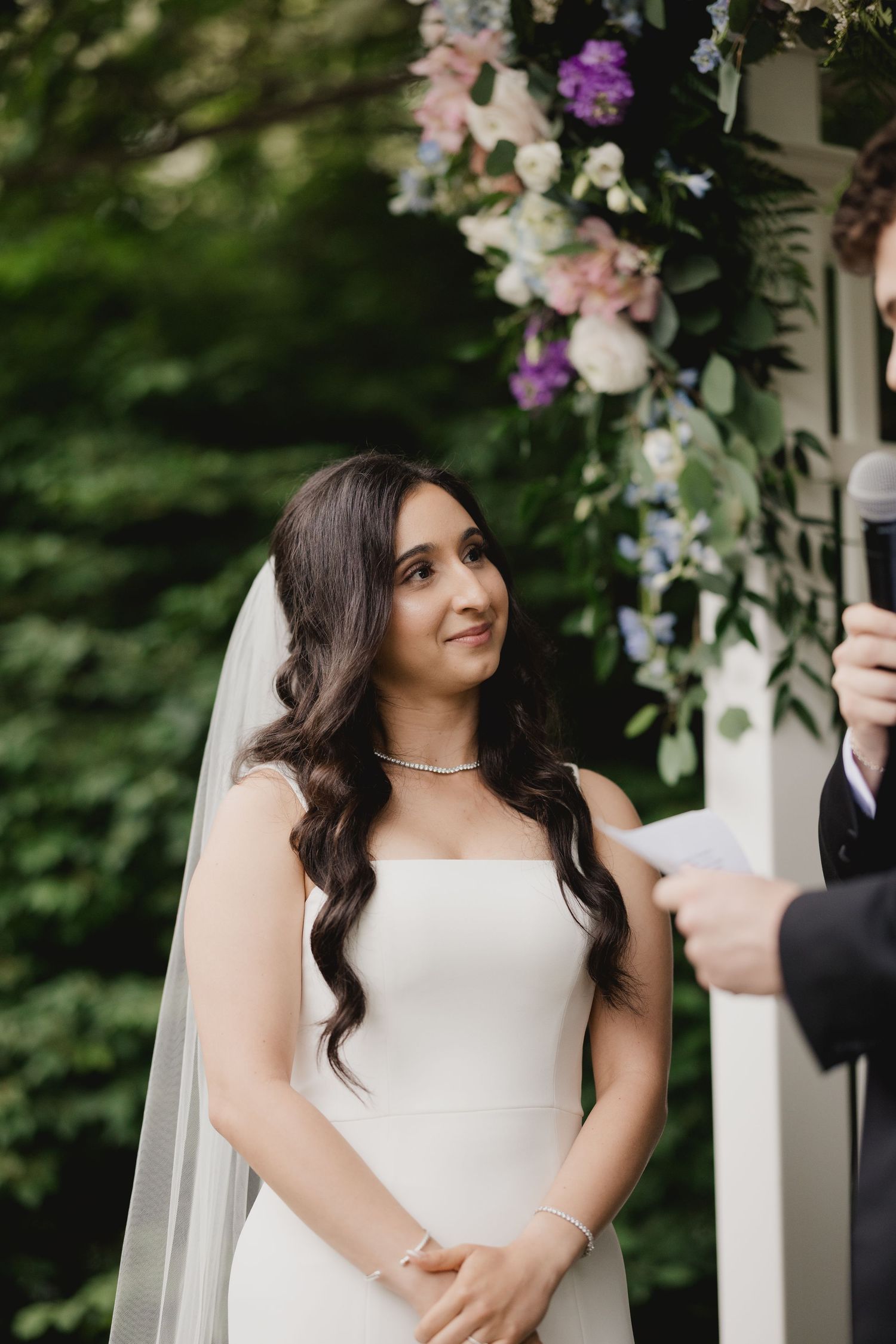 Bride in strapless white gown and veil smiles during outdoor wedding ceremony beneath floral arch.