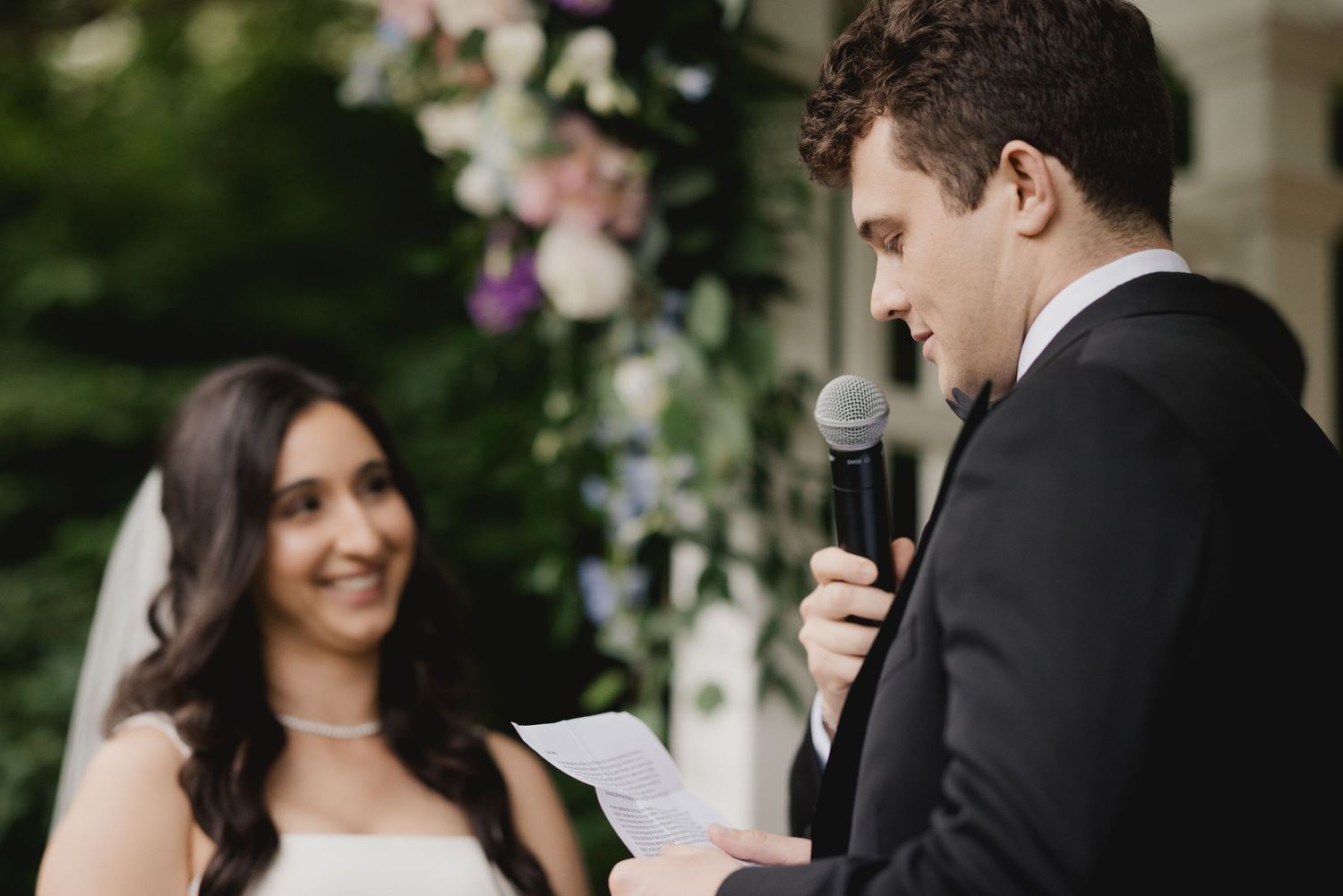 Groom reads vows from paper at outdoor ceremony while bride smiles beside floral arch backdrop.