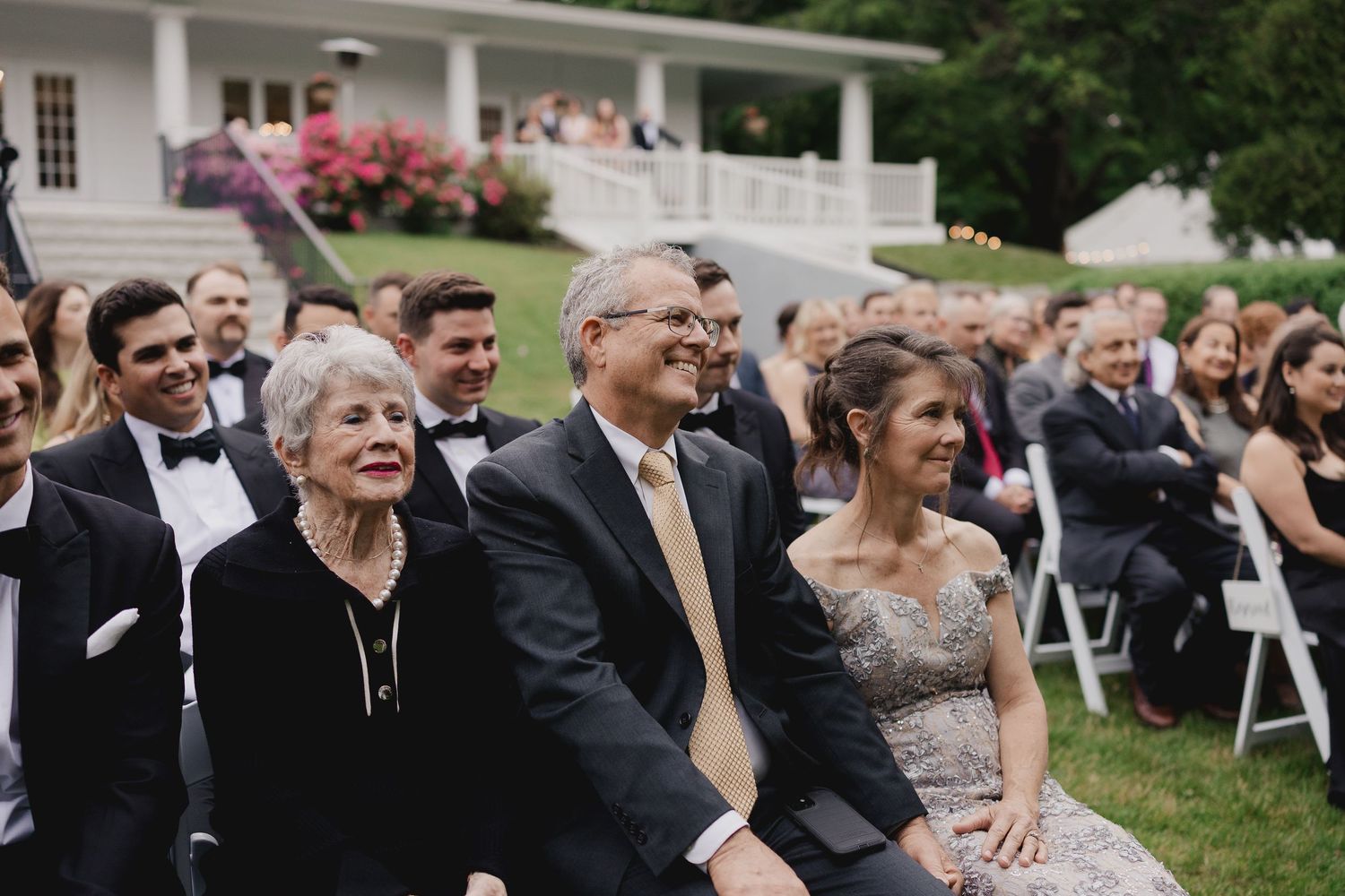 Smiling wedding guests seated outdoors at elegant ceremony with lush garden backdrop at estate venue.
