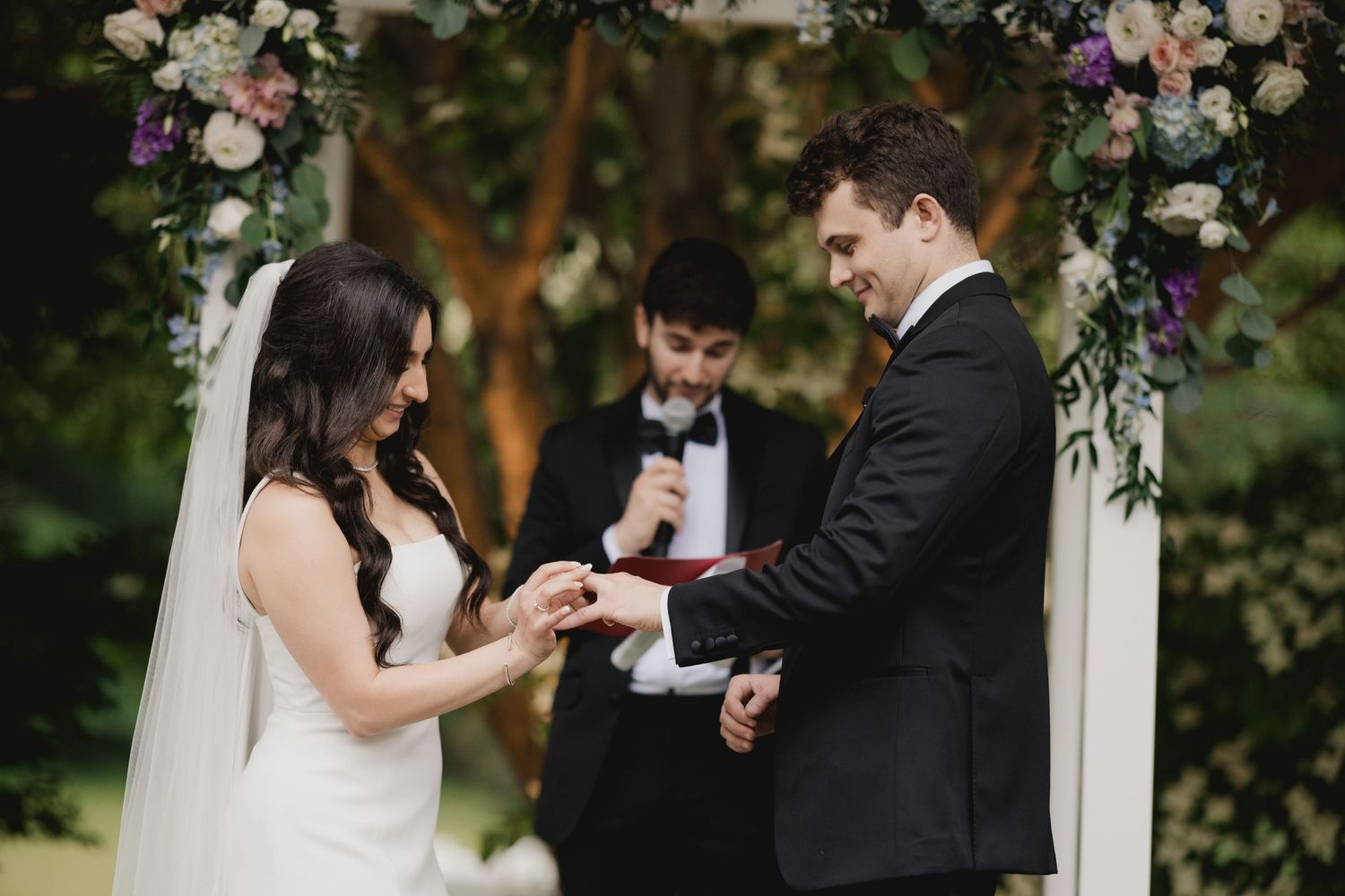 Bride and groom exchanging rings during outdoor wedding ceremony with floral arch and officiant.