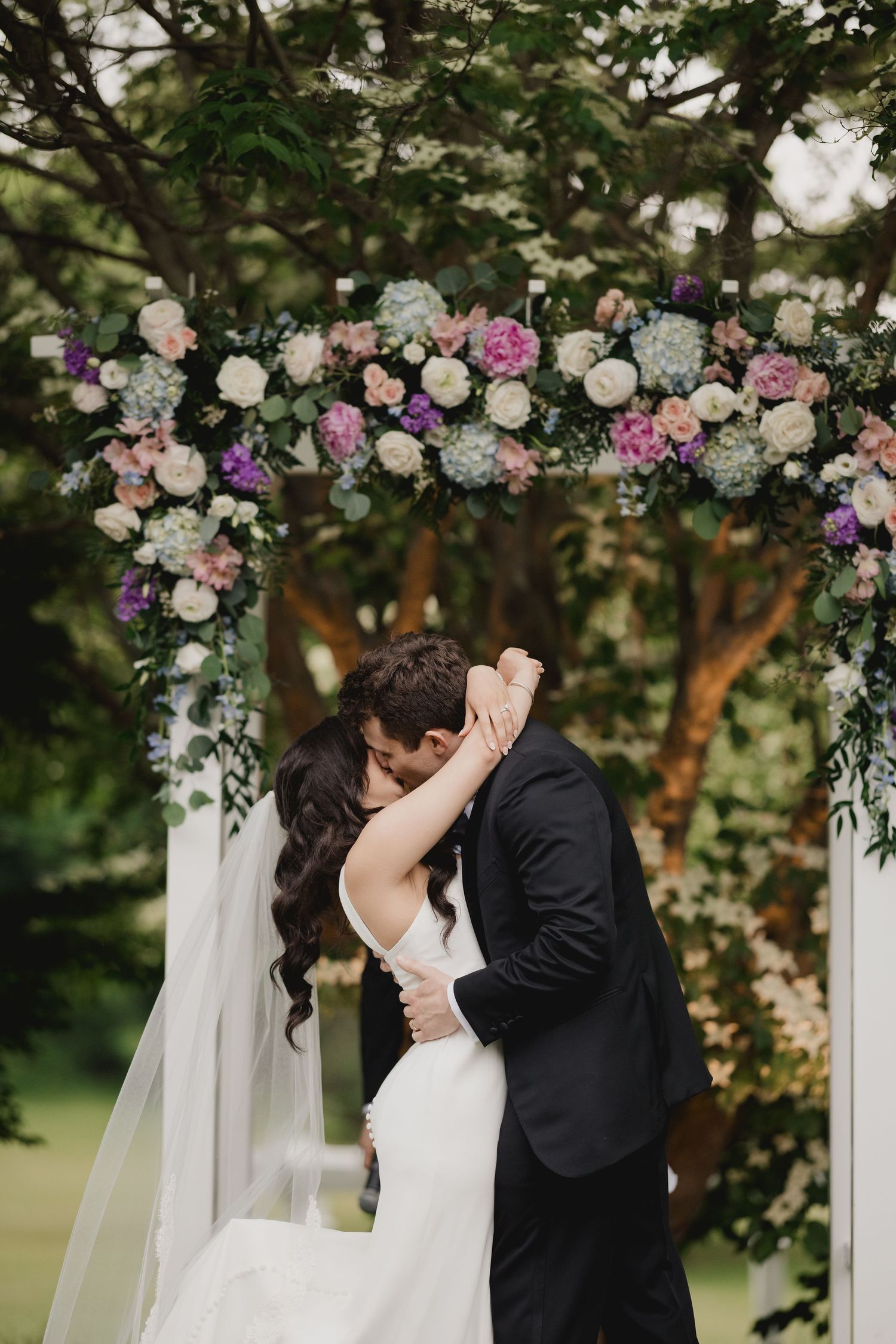 Bride and groom share first kiss under floral wedding arch with pink and white blooms.
