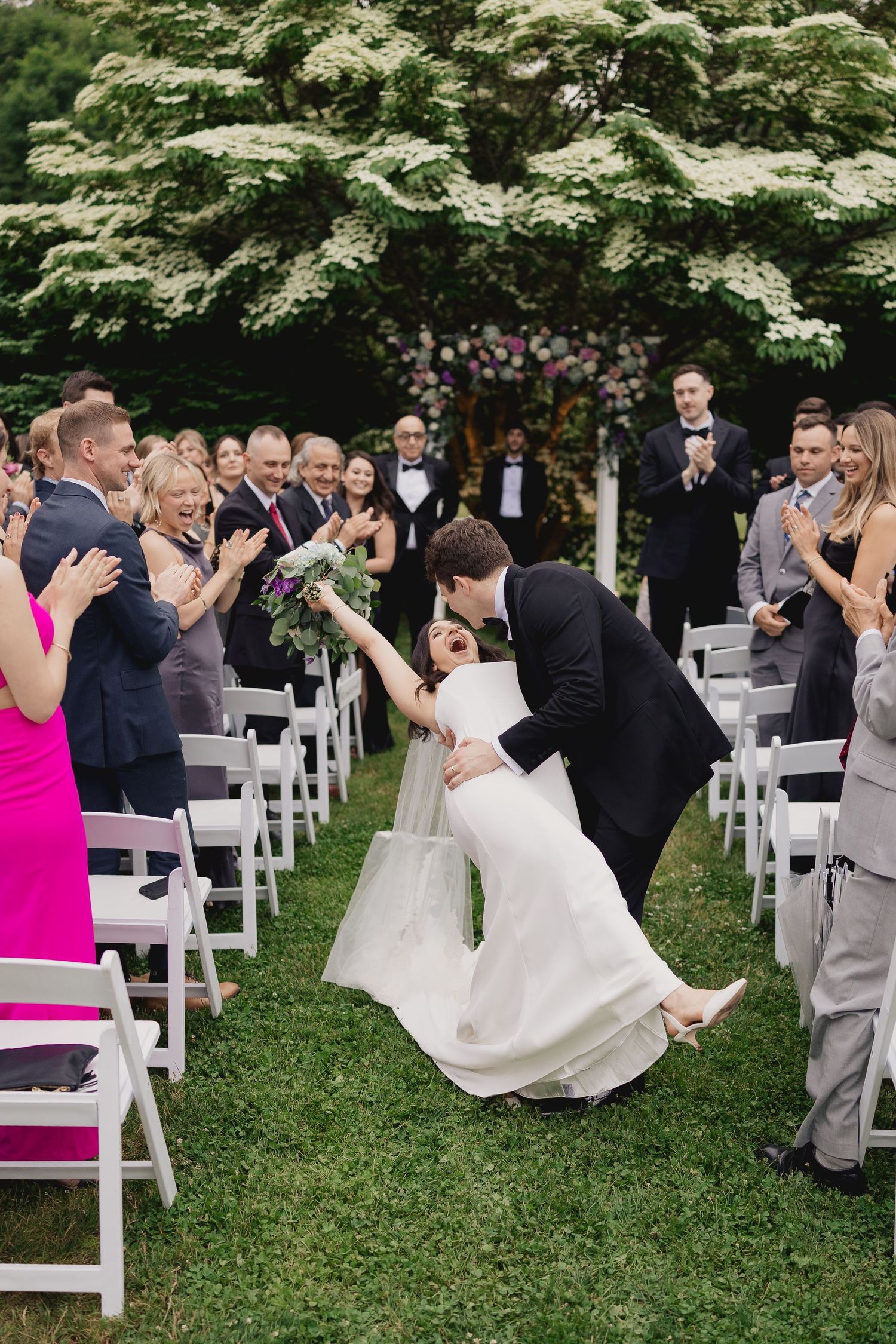 Groom dips bride for a kiss in the aisle as guests cheer at outdoor garden wedding ceremony.