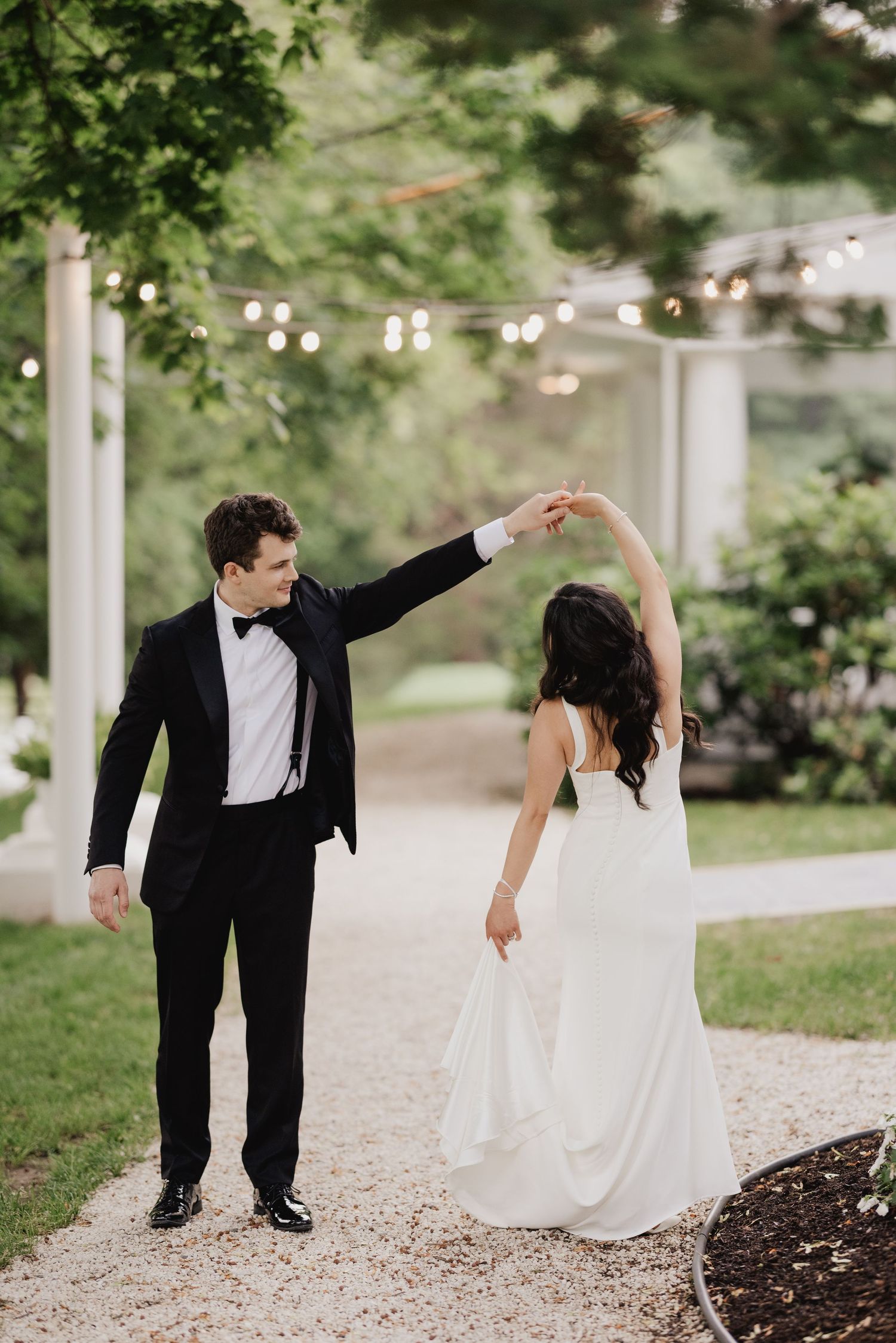 Groom in black tuxedo spinning bride in white wedding gown on garden path with string lights.