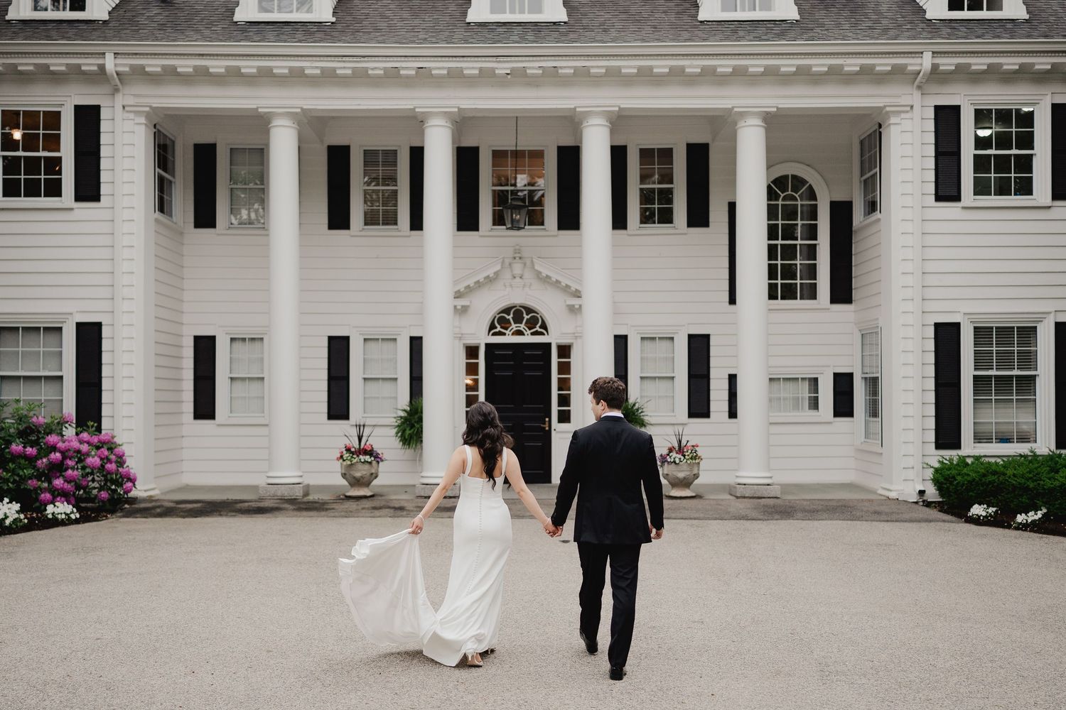 Bride and groom walk hand in hand toward grand white colonial wedding venue with tall columns.
