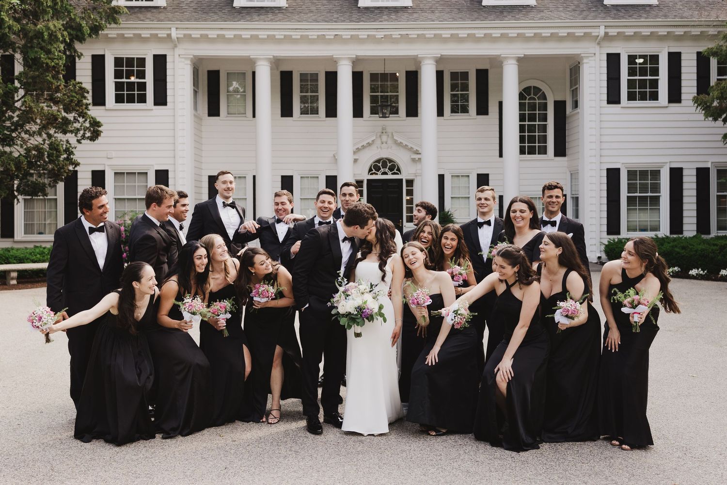Wedding party in matching black attire poses with bride and groom outside white colonial mansion.
