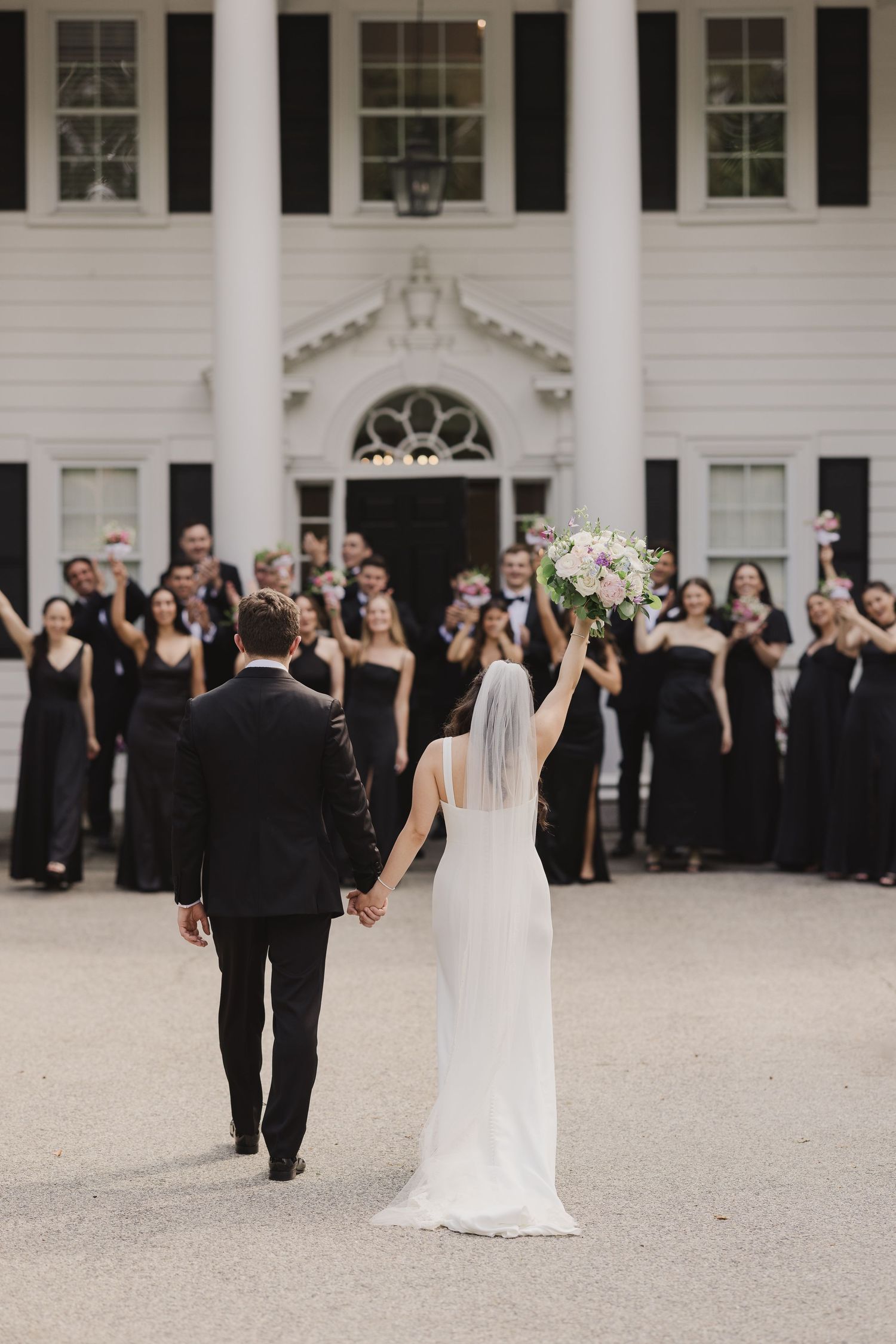 Bride raises bouquet as couple walks toward wedding party in black attire outside elegant white colonial mansion.