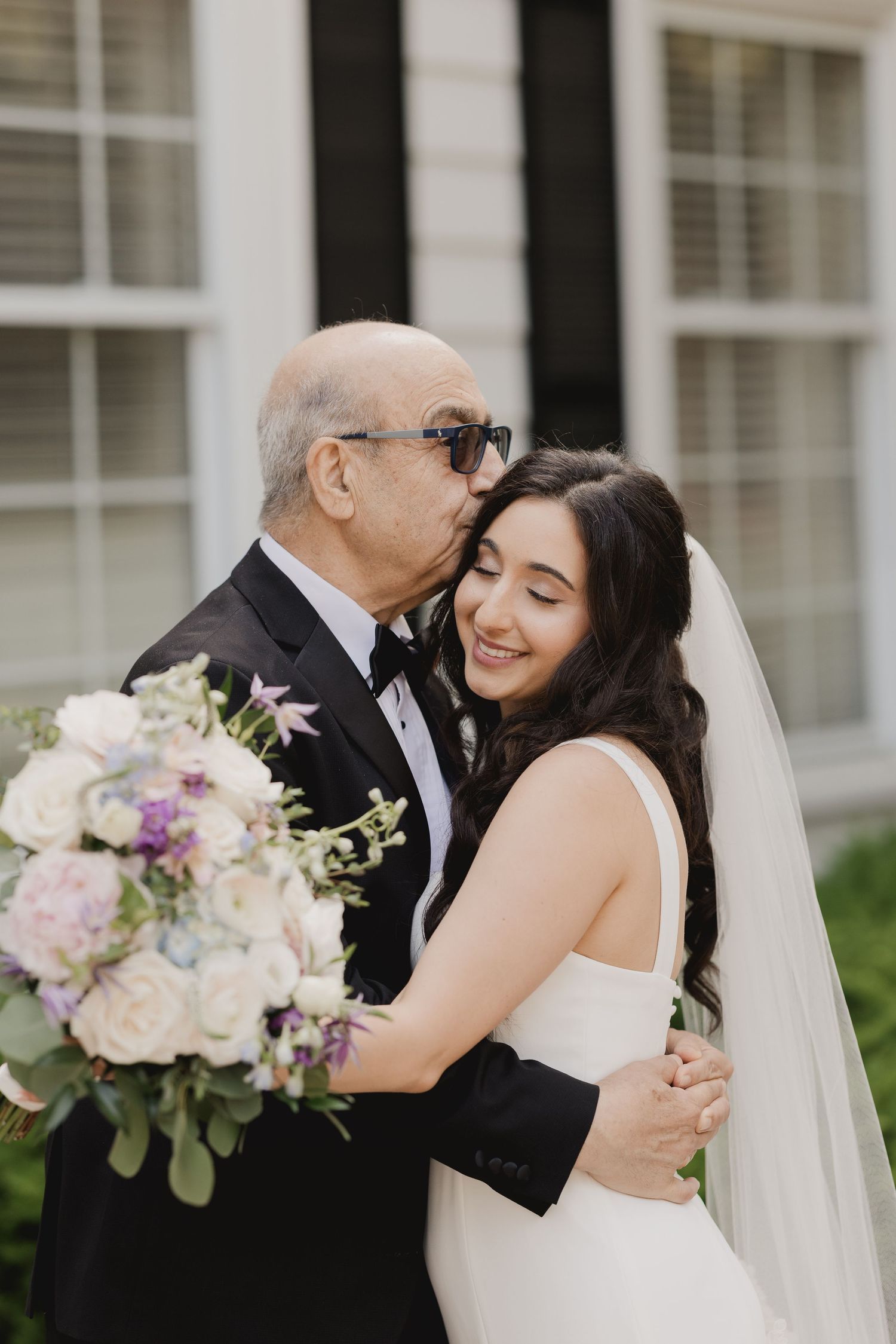 Father kisses bride on cheek as she smiles holding lush pastel bouquet outside wedding venue.