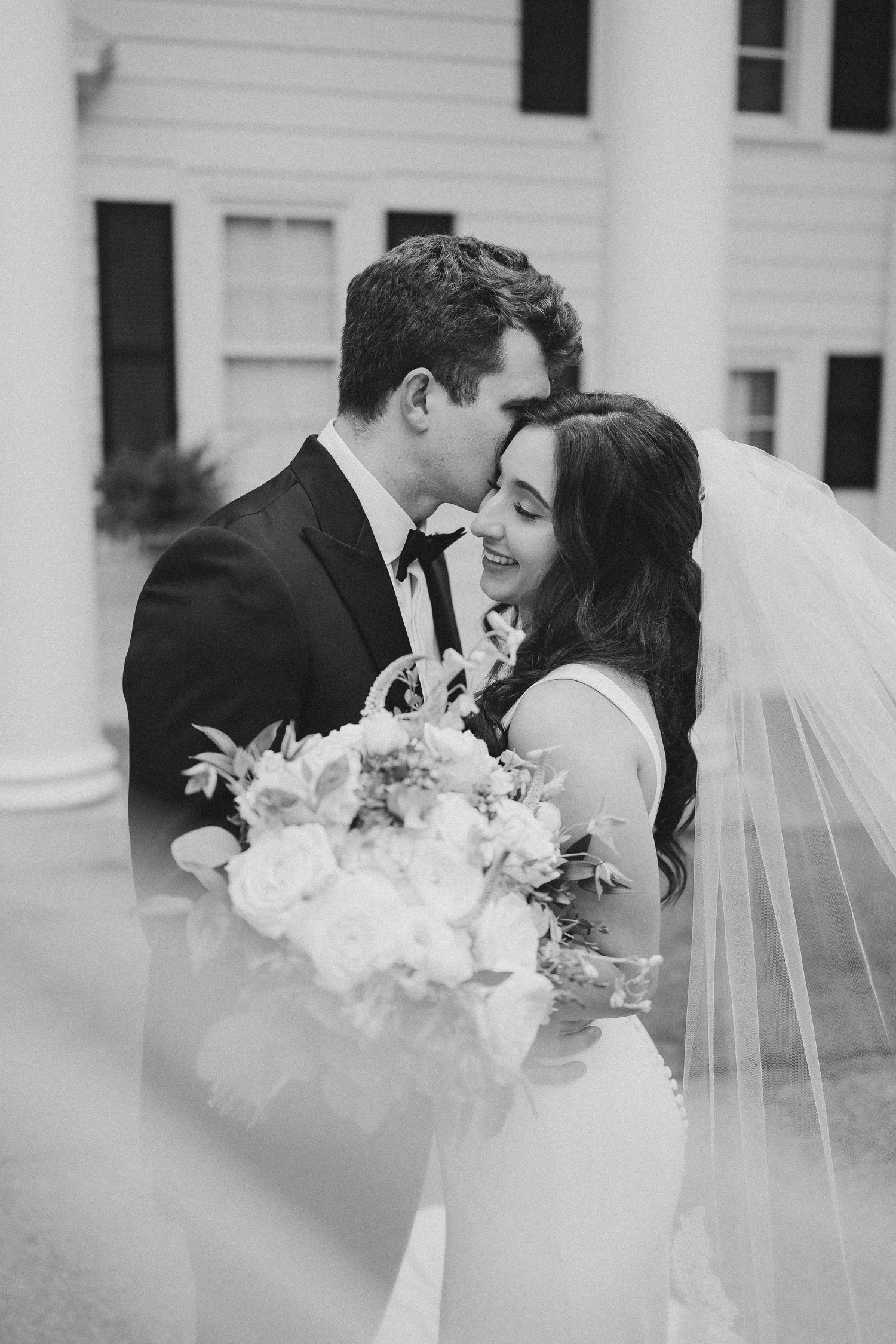 Black and white portrait of groom kissing bride's cheek as she smiles holding floral bouquet.