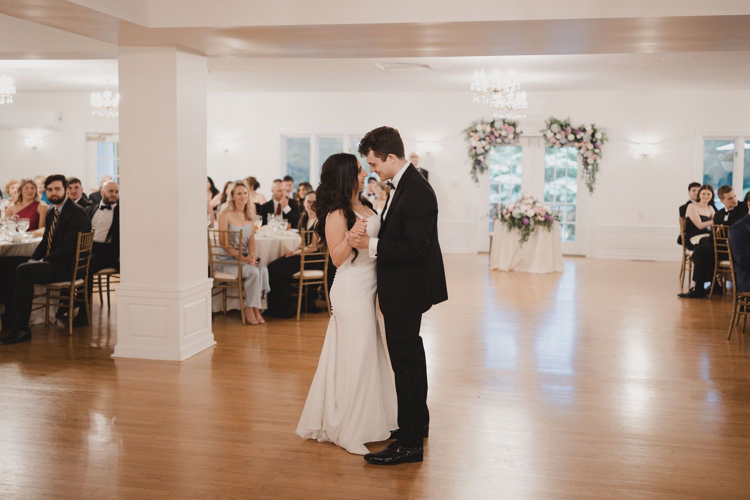 Couple shares their first dance on a wooden floor in an elegant white ballroom with floral arch and seated wedding guests.