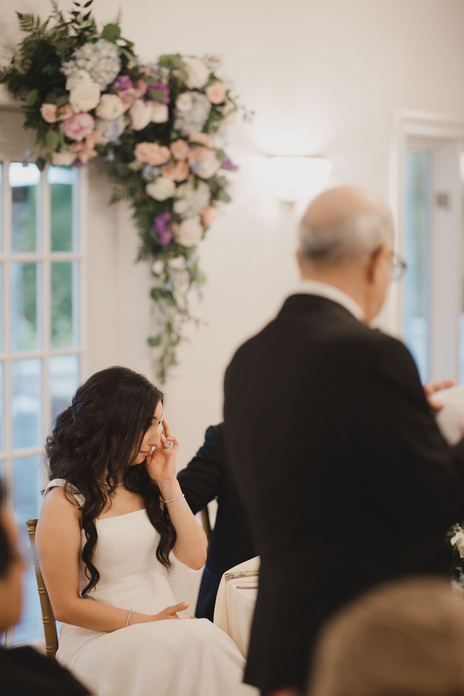 Emotional bride in white gown wiping tears during heartfelt wedding reception speech.