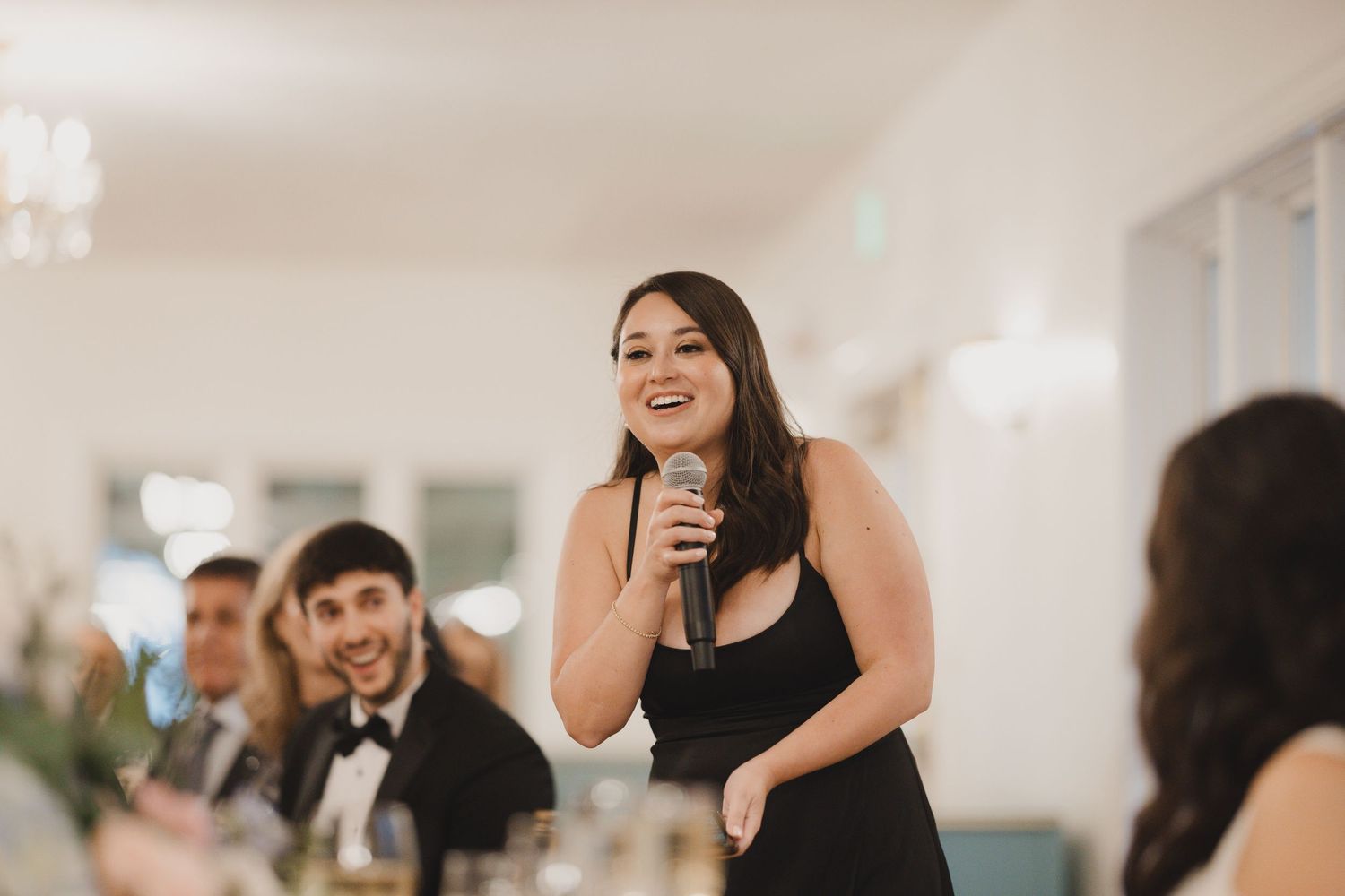 Woman in black dress gives speech holding microphone at elegant wedding reception.