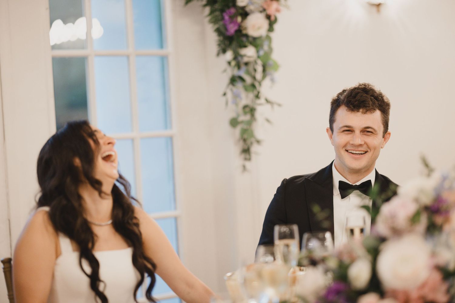 Bride and groom laughing at wedding reception table with floral arrangements.
