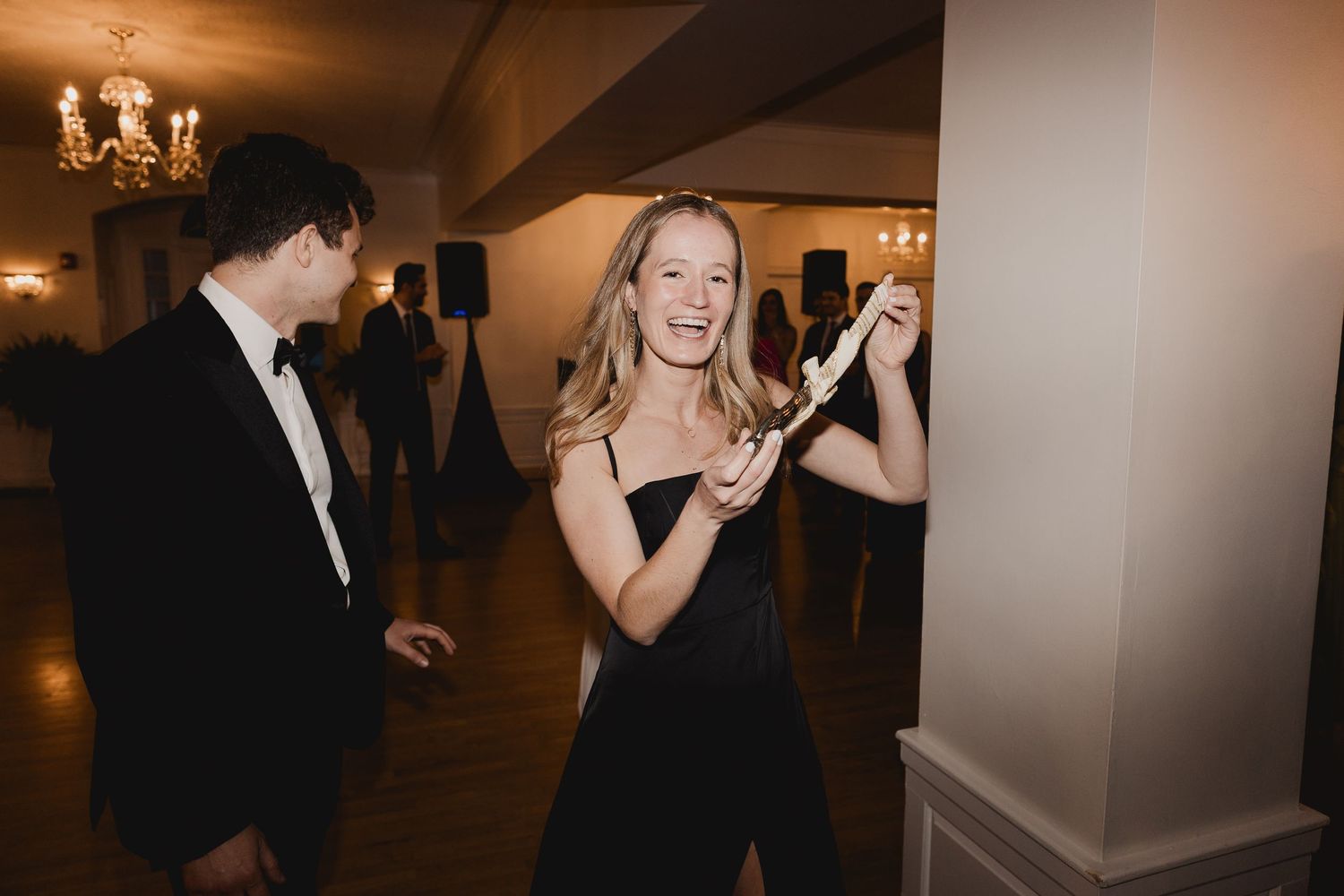 Woman in black dress smiling and dancing at elegant formal event with chandelier lighting.