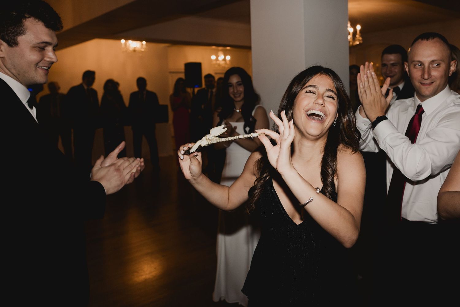 Woman laughing joyfully on wedding reception dance floor surrounded by clapping guests in formal attire.