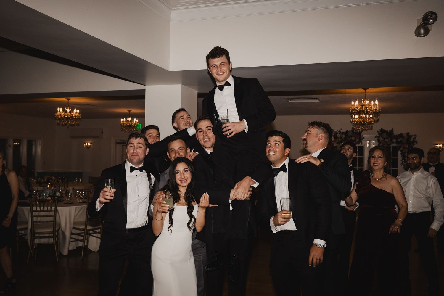Groomsmen in tuxedos lift groom on shoulders during lively wedding reception celebration.