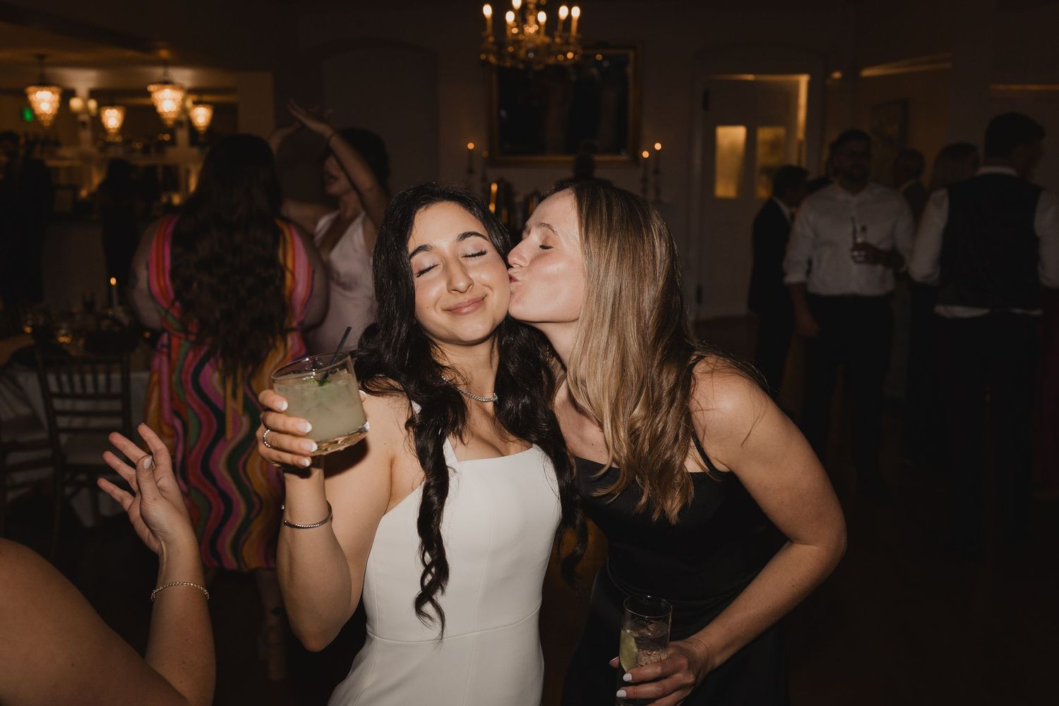 Two women celebrating at a wedding reception, one in white dress receiving a kiss on the cheek while holding a cocktail.