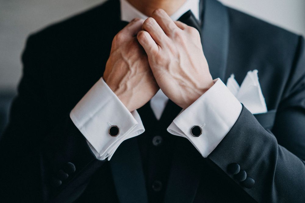 Close-up of formal black suit with hands adjusting white tie and cufflinks.