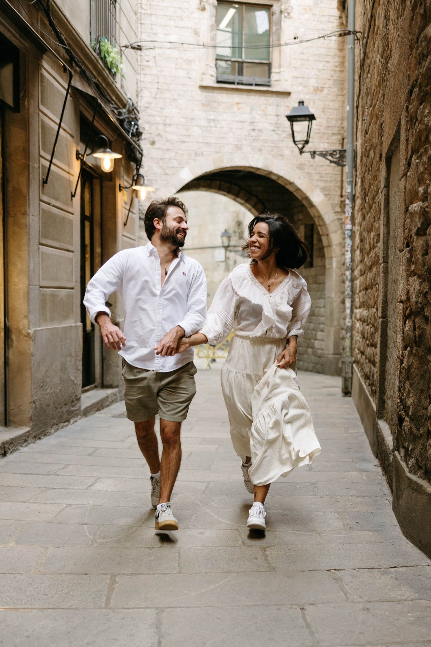A carefree couple running through a charming stone archway in coordinated white outfits.