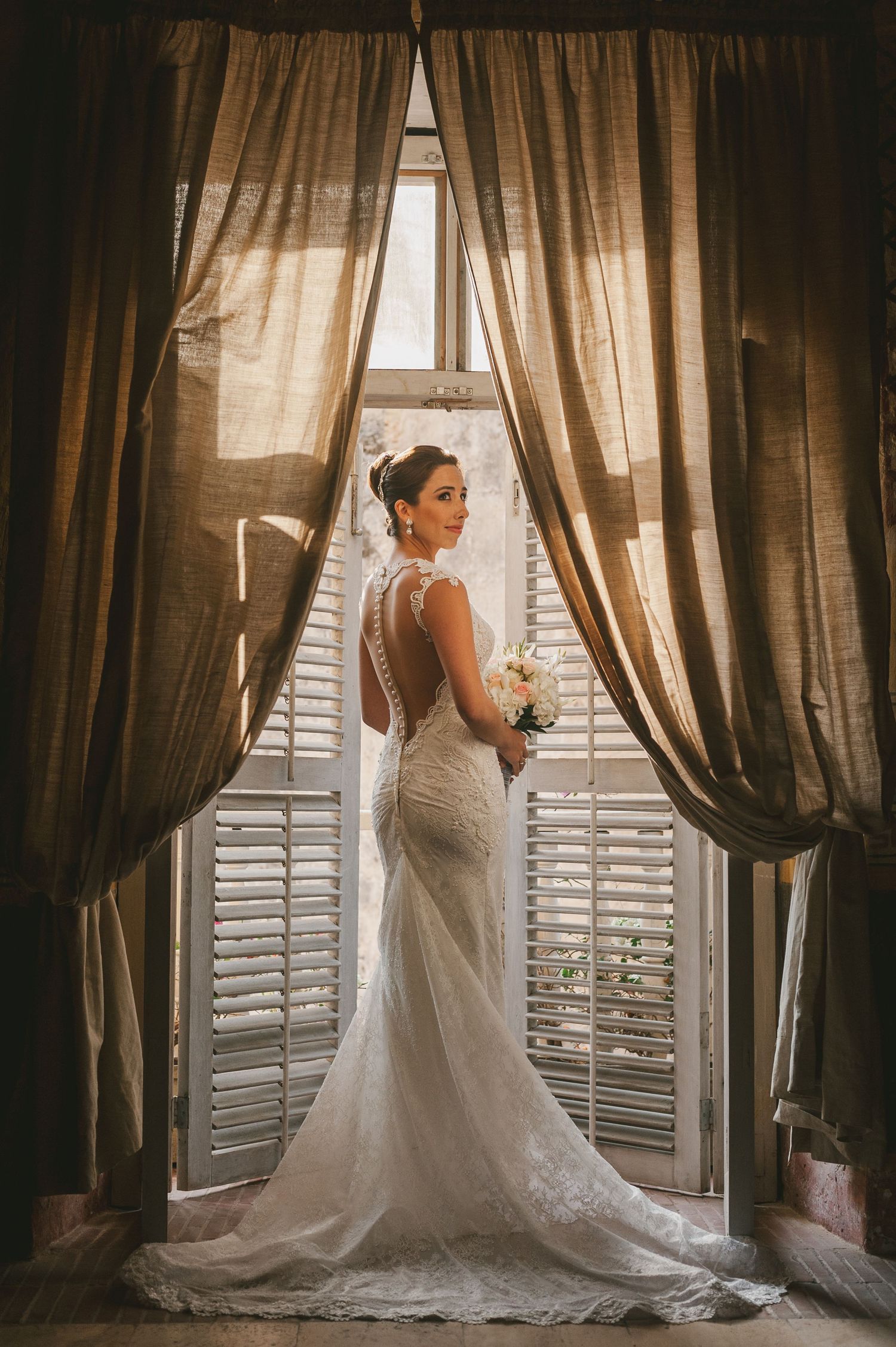 A bride in an elegant white dress sits atop a brown horse decorated with white ribbons near lush magnolia trees.